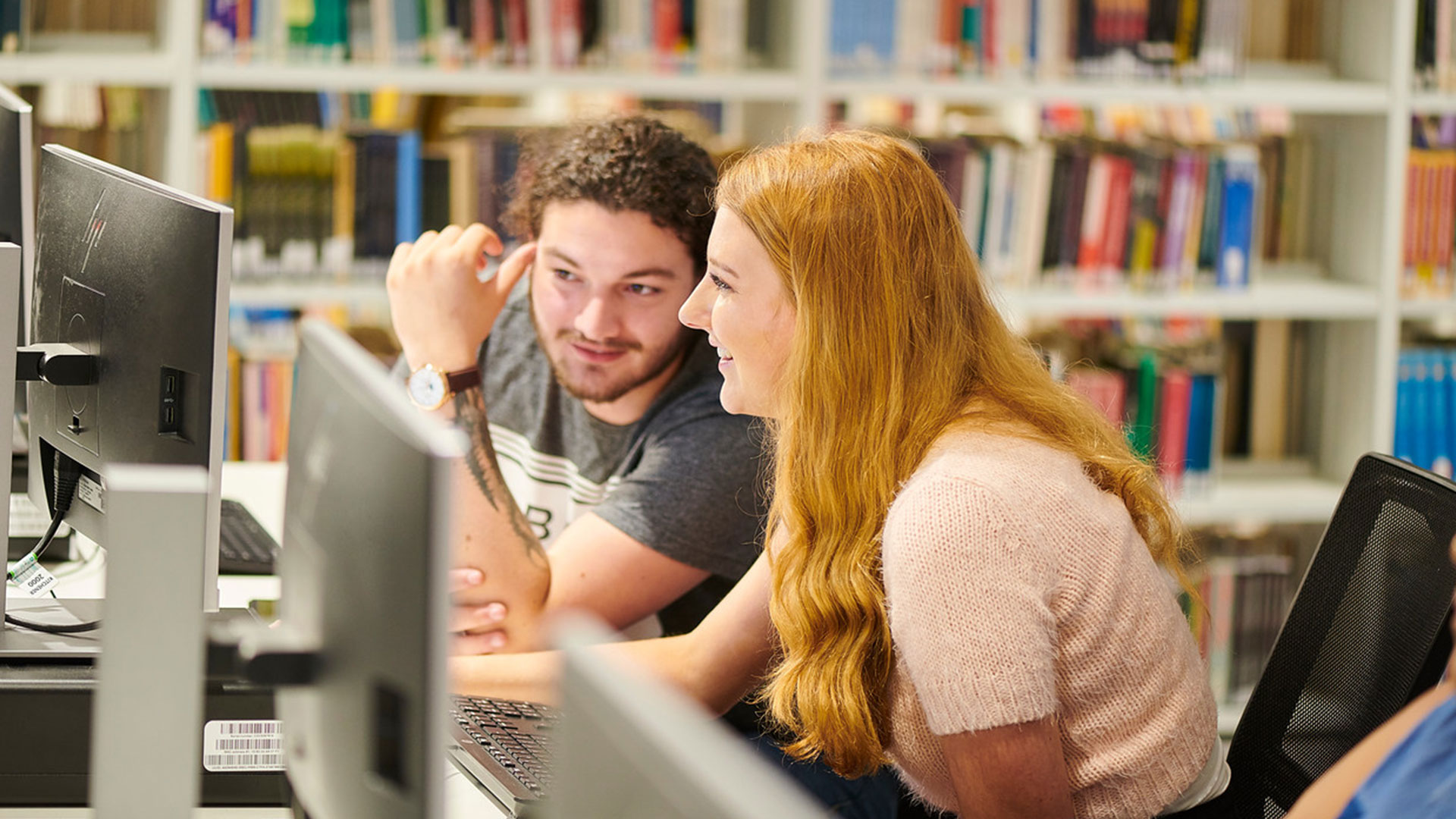 Two students in the library with rows of books in the background, looking at a computer screen together