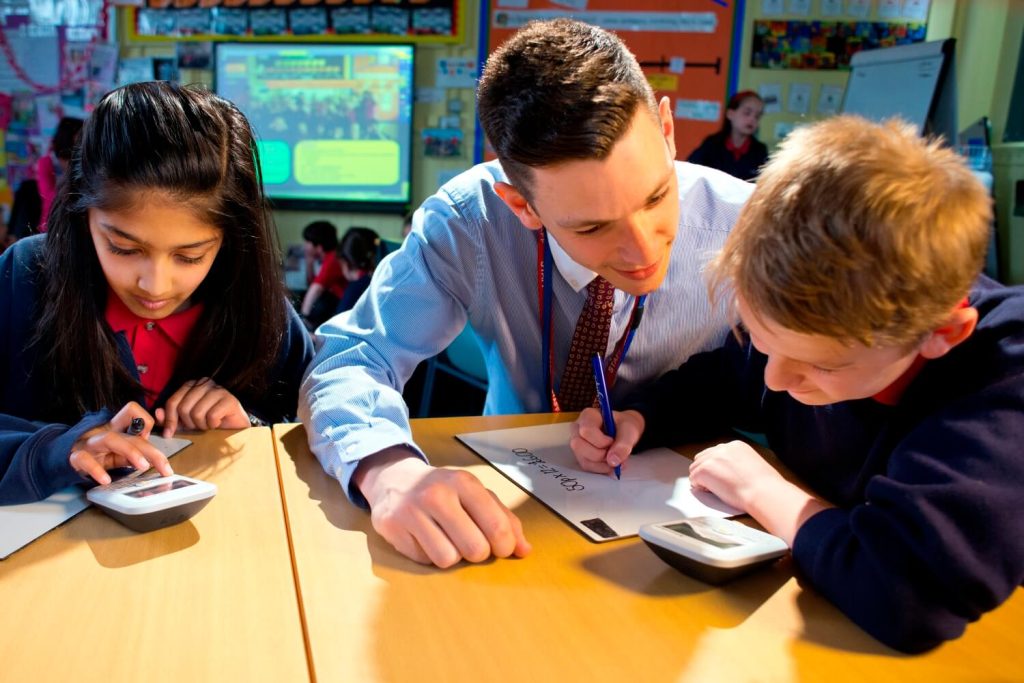 A teacher observes as two pupils use calculators during a maths lesson in a secondary school classroom.