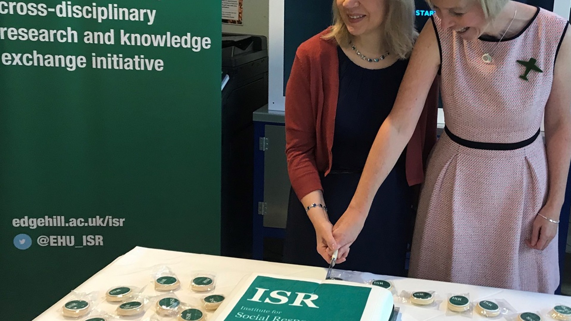 Two people are stood cutting into a cake that says ISR on a green background. There is a green sign next to them that describes ISR on a green background.