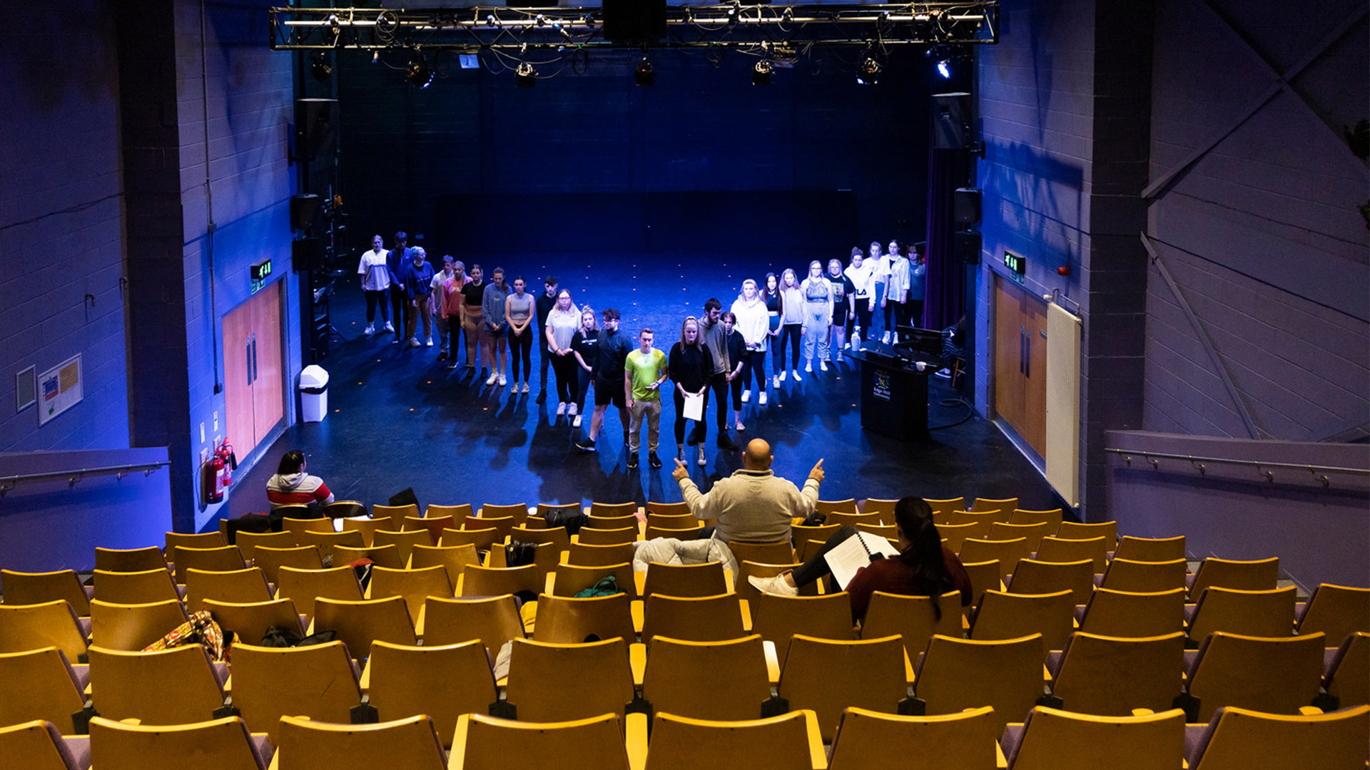Anempty theatre auditorium with rows of empty seats and students rehearsing on stage