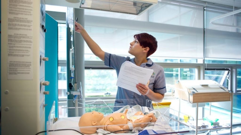 A nurse is looking at a fluid bag, which is attached to a baby practice mannequin that is at the forefront of the image.