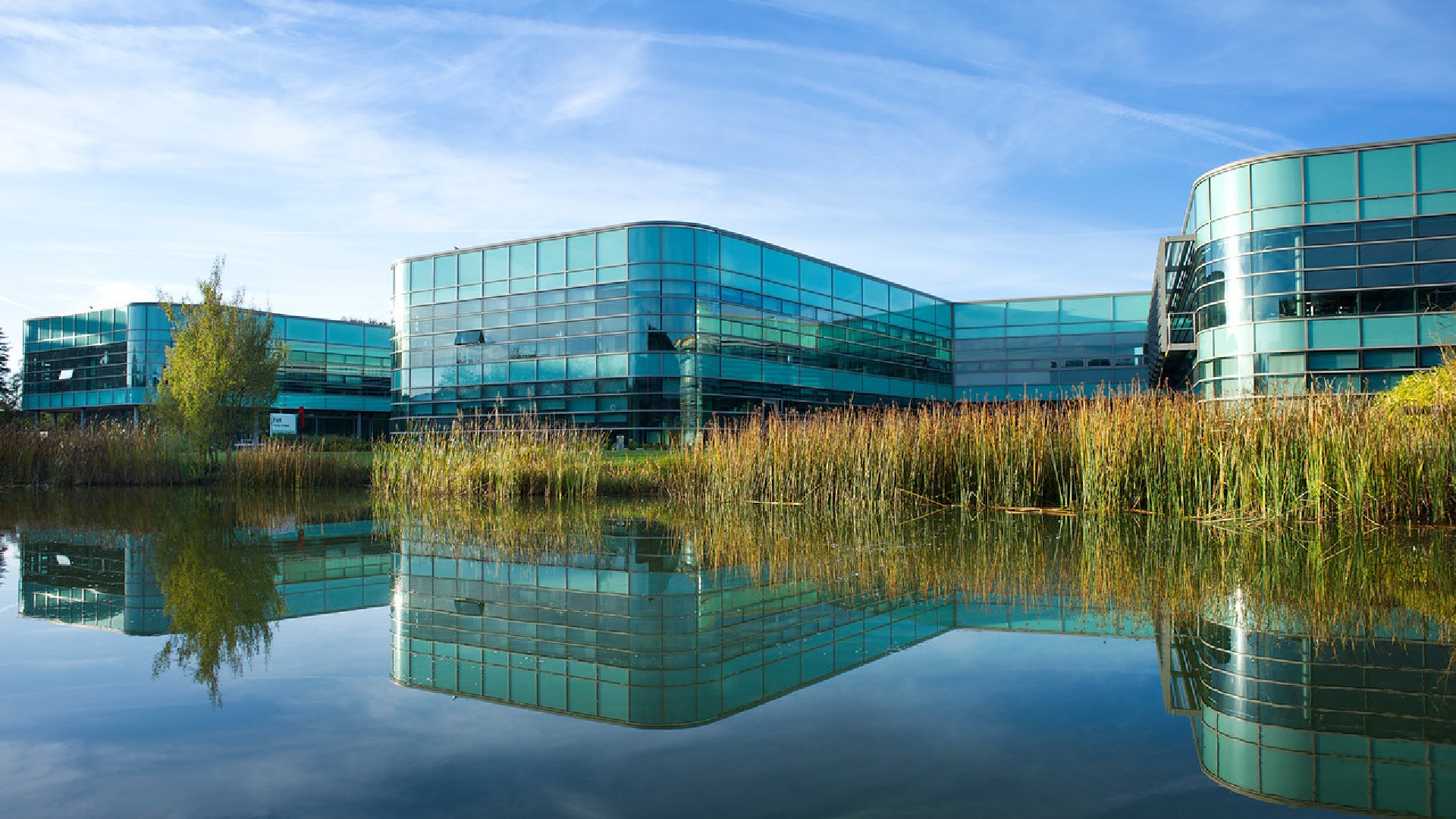 An image of the health department building at Edge Hill, there is a tall glass building in the image, in the foreground of the image there is a lake which has a reflection of the building on.