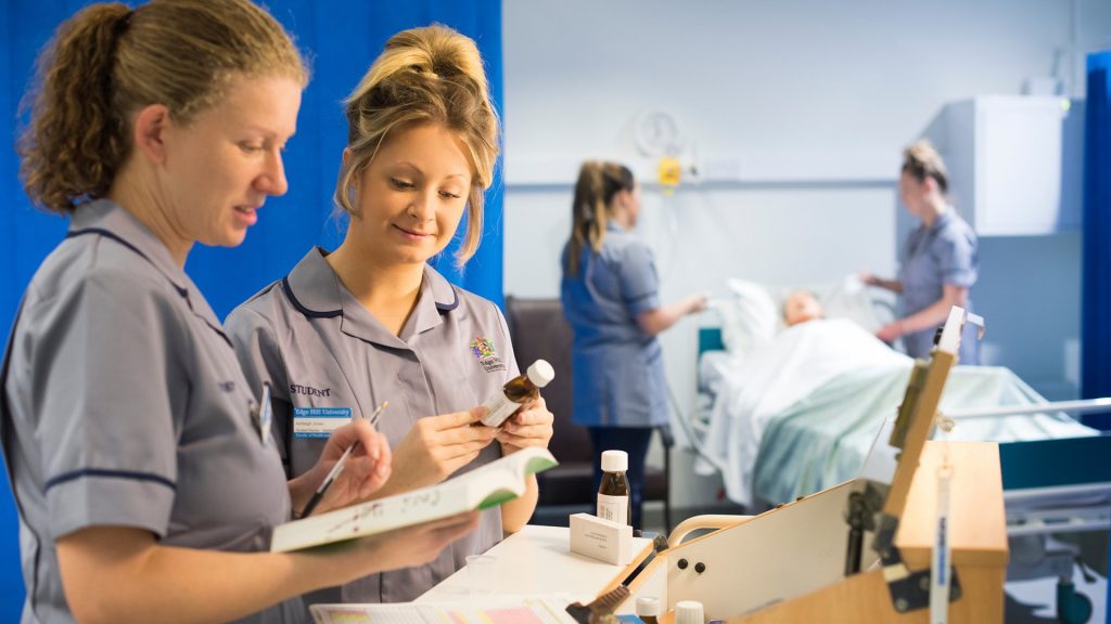 Two student nurses are stood looking at a booklet. One is holding a pen, the other is holding a bottle of medicine. There are various forms of medicines in front of them on a table. There are two other nurses in the background of the photo interacting with a practice mannequin.