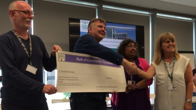 Four people are holding a cheque that has the text "Bank of Community Champions" on, they are all looking away from the camera and smiling.