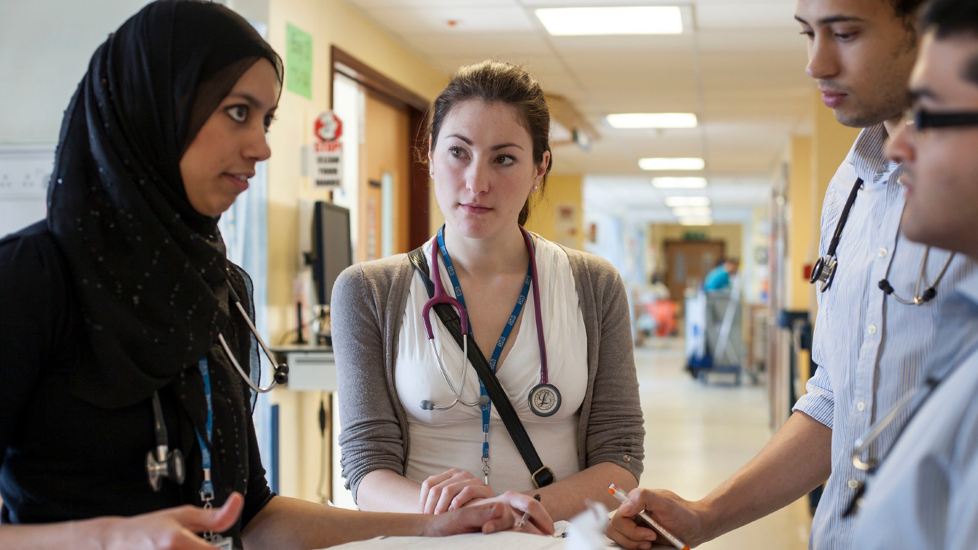 4 people are stood around a stack of paper, they are stood inside a well-lit corridor. All people in the photo are wearing stethoscopes.