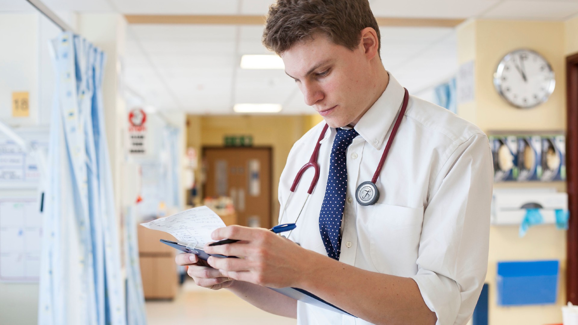 A person is stood looking at paper on a clipboard. They are wearing a stethoscope.