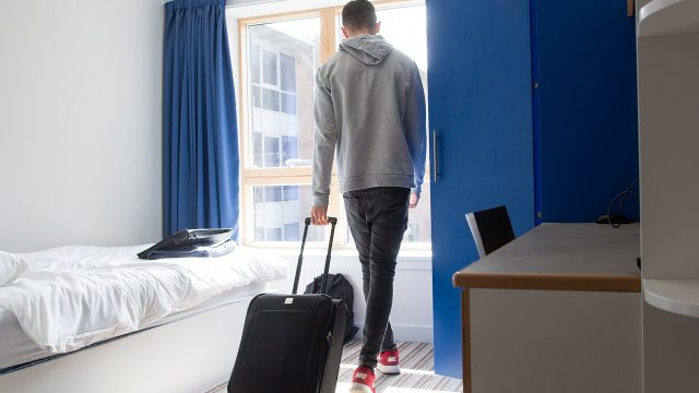 An image of a person walking into an accommodation room at Edge Hill university. They are carrying a black suitcase and walking towards the window of the room. 
