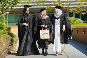 Three graduates, one carrying an alumni bag, walk together on the western side of campus after their graduation ceremony.