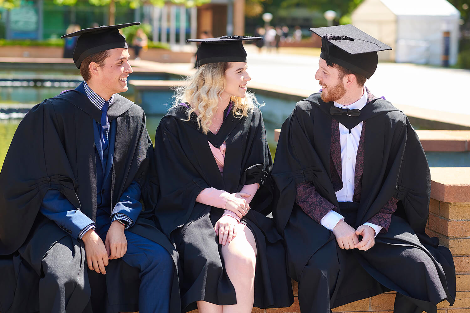 Three graduands sit in the piazza area on the western side of campus while wearing their graduation robes and mortar boards.