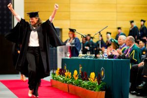 A student holds her arms aloft as she walks along the red carpet during a graduation ceremony.
