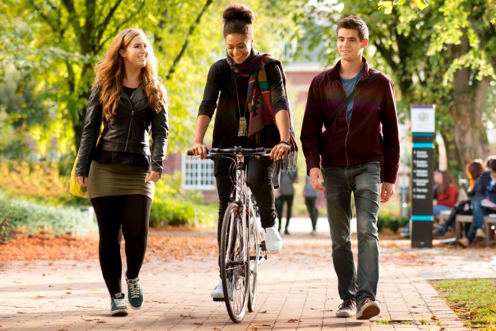 Three students, one of them on a bicycle, near the Business School and Law & Psychology building.