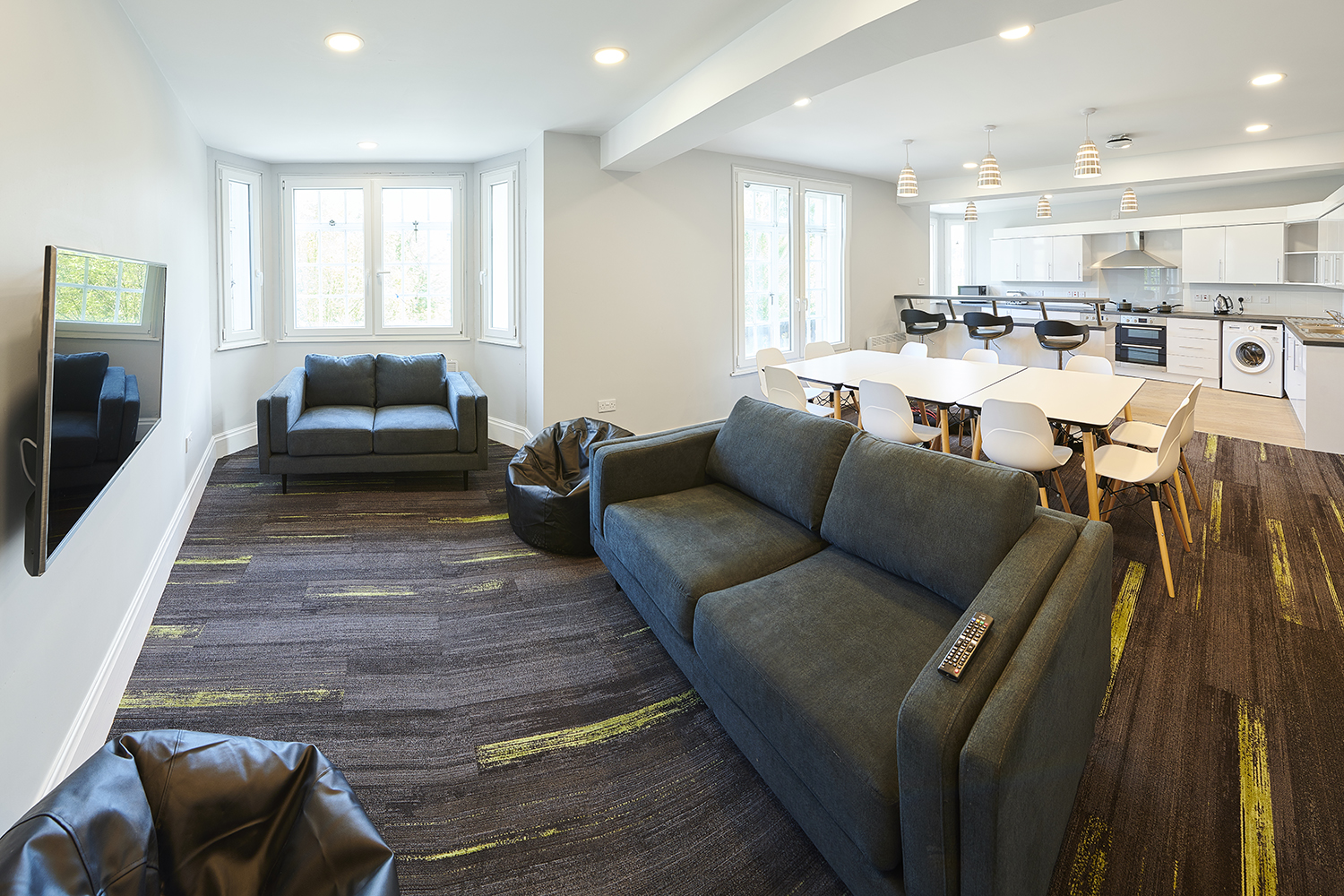 Lounge area in a set of flats in the eastern wing of Main Halls, with sofas and bean bags arranged around a flatscreen TV on the wall, with a dining area, breakfast bar and kitchen visible in the background.