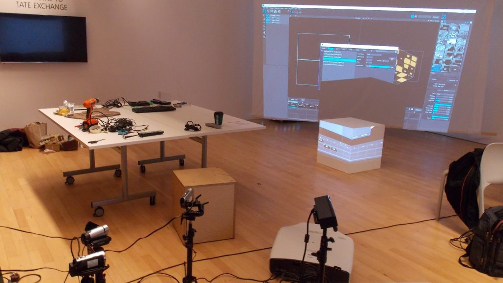 An image of an exhibition at the Tate Liverpool. There is a table and multiple pieces of equipment in this image and a projector that is showing a display on one of the walls.