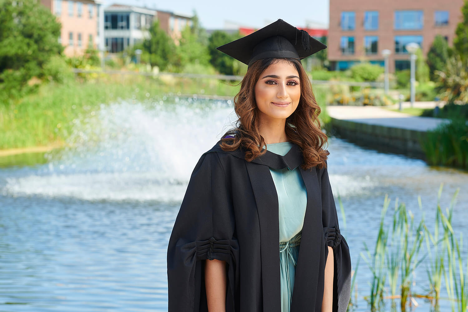 A graduate poses for a photo in her robe and mortar board by the lake on the eastern side of campus.
