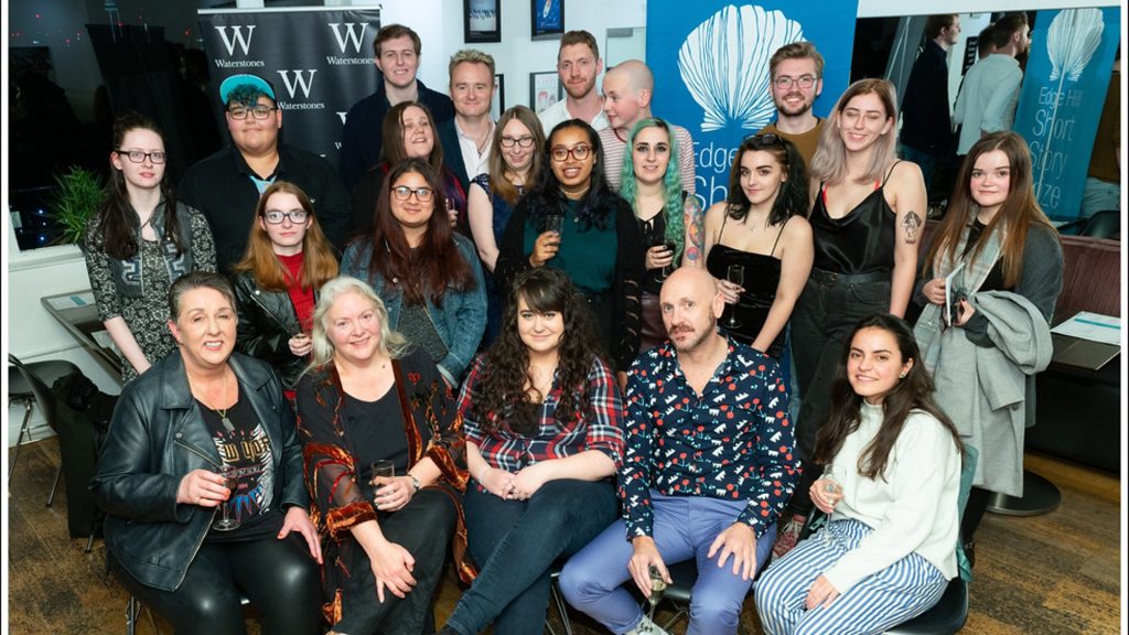 An image of a group of students stood in front of two banners, one has the waterstones logo on a black background, the other says "Edge Hill, Short Story Prize"