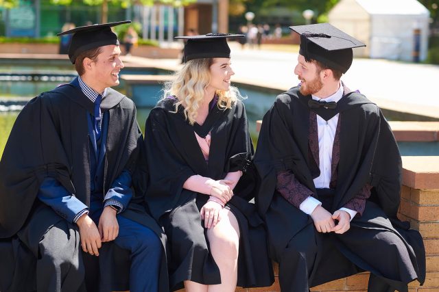 Three graduates sit on the piazza wearing their caps and gowns.
