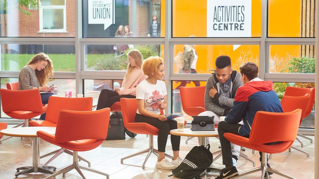 An image of multiple people sat around various small tables in The Hub on the Edge Hill University campus. One set of these groups of people are looking at an iPad that is placed on the table.