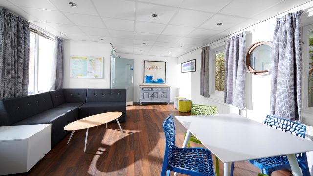 An image of one of the blocks of accomodation at Edge Hill university. The image features the Margaret Bain block within the back halls accomodation. The image highlights one of the common rooms in the accommodation, there are various seating areas and large tables in a white room that has multiple windows.