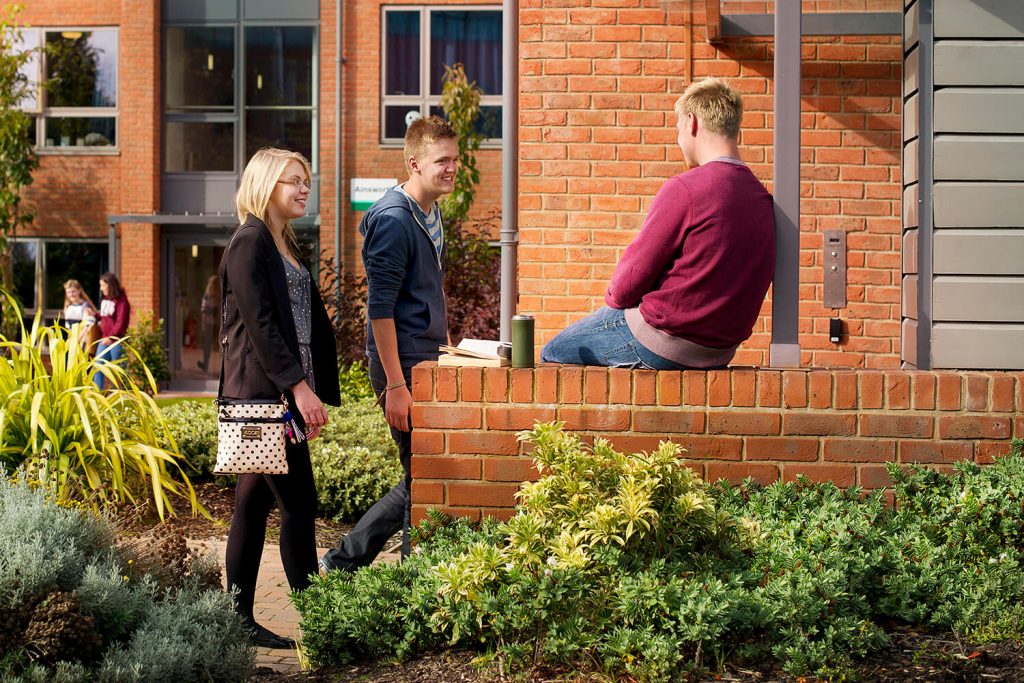 Three students, one sat on a wall, outside the entrance to a hall in Graduates Court.