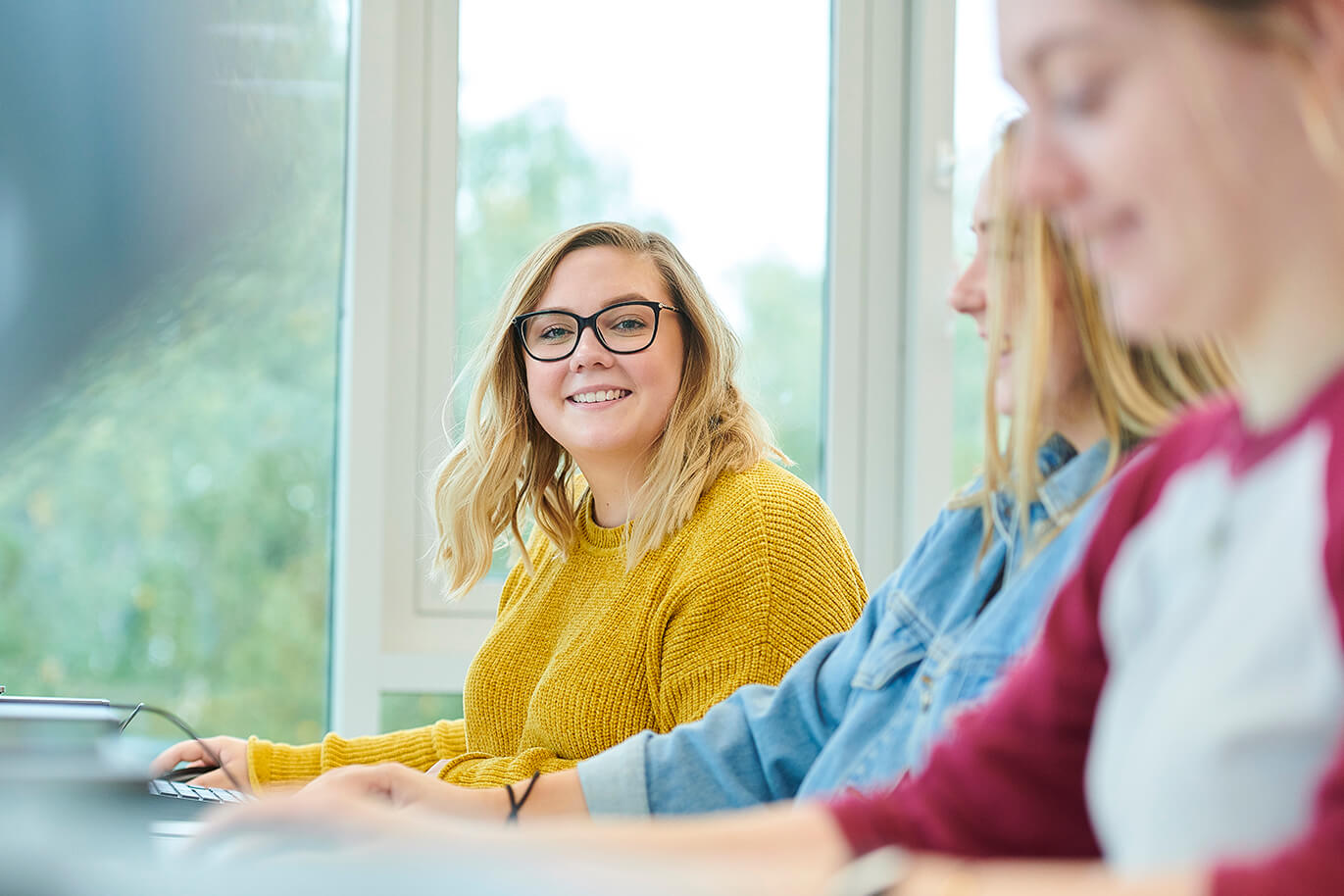 Three students sit at desks in front of computer screens.