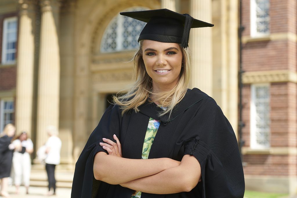 A graduate stands in her robe and mortar board with arms folded outside the front entrance to the Main Building on campus.