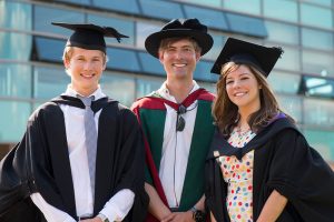 Three graduates stand outside the Faculty of Health, Social Care and Medicine