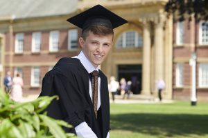 A graduate poses for a photo in his robe and mortar board at the front of the Main Building on campus.