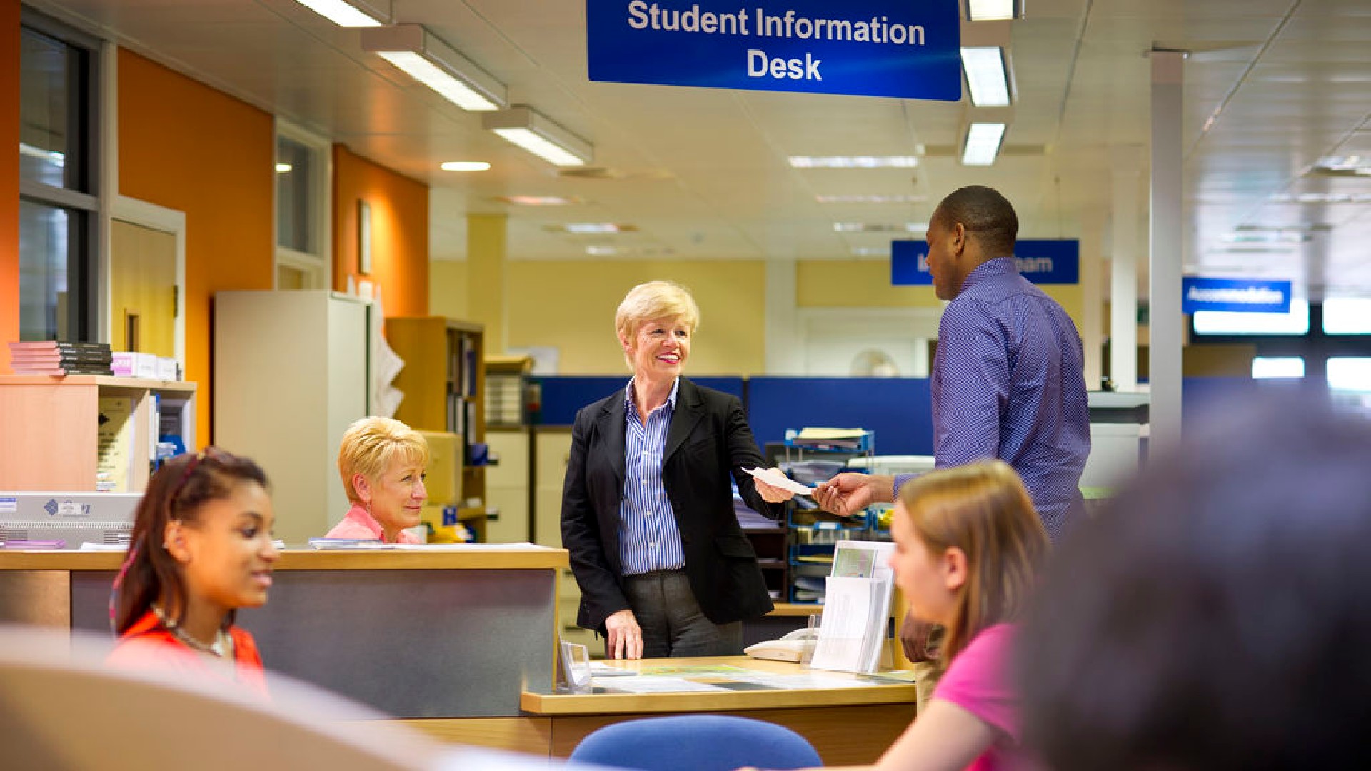 A group of people are stood near a desk that has the sign "Student Information Desk", in front of the desk two people are sat talking. 