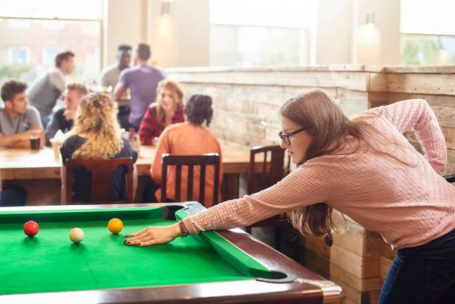 A student plays pool in the SU Bar on campus. Other students can be seen enjoying a meal and drinks.