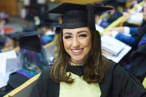 A graduate sitting in a lecture theatre in the Faculty of Health, Social Care and Medicine with their fellow graduands as the ceremony is about to begin.