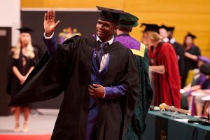 A graduand waves to a guest in the audience after walking along the red carpet to shake the hand of Dr John Cater during a graduation ceremony.