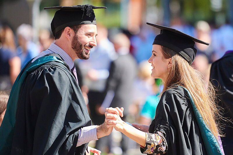 Two graduates hold hands while stood in their robes and mortar boards outside on the western side of campus.