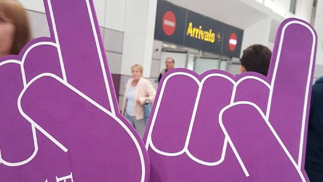 An image of two purple hands that have white text on that says "Edge Hill University", in front of an airports arrivals corridor. 
