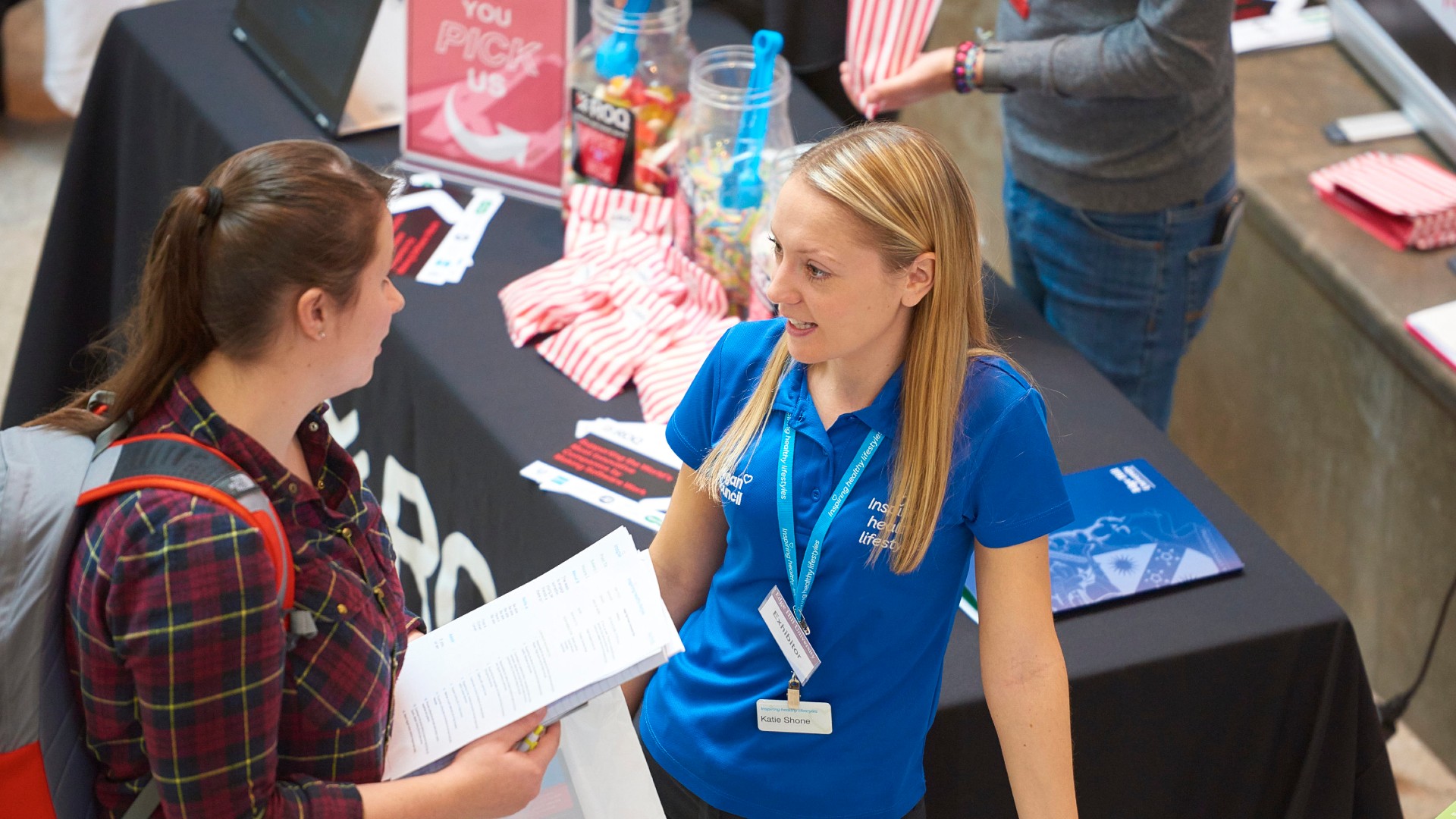 Two people are stood next to a table at a careers fair. 