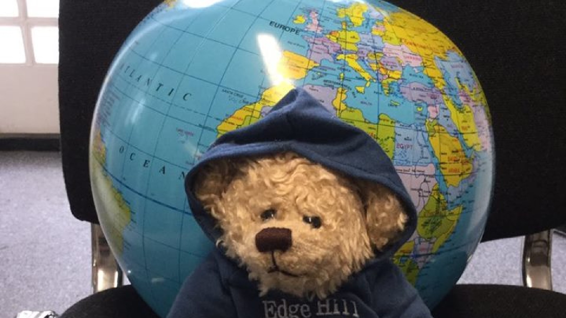 An image of a small brown bear wearing a blue jumper that has white text on that says "Edge Hill University". Behind the bear is a globe. Both the bear and the globe are on top of a black chair. 