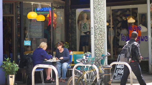 Two people are sat on a small table outside a cafe, a person is walking past them. 