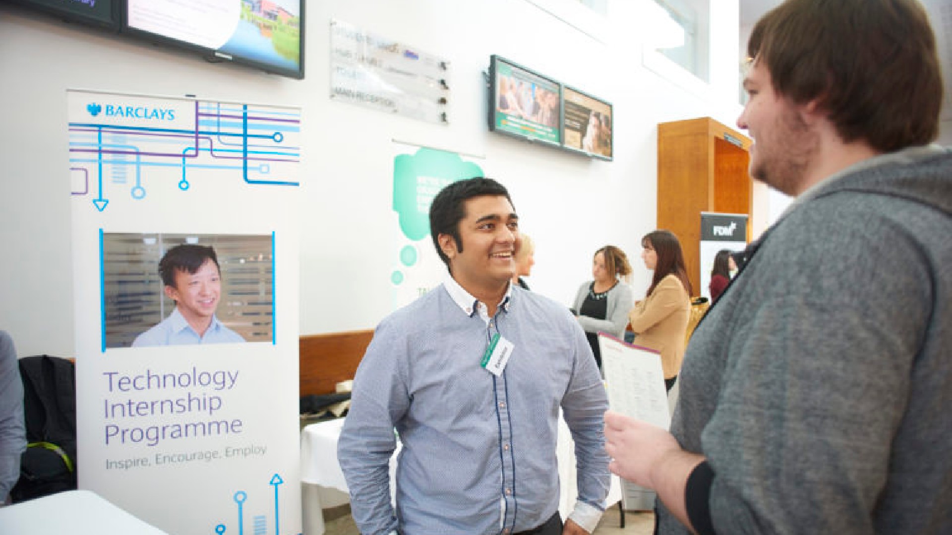 A student is talking to an employer in front of a sign that says "Barclays, Technology Internship Programme"