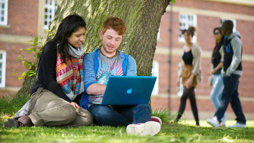 Two students sat outside looking at a laptop and laughing