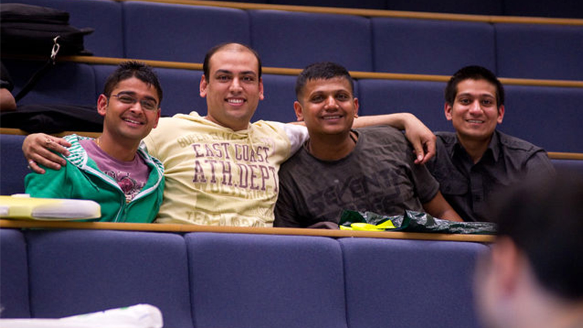 Four students sit in a row in a lecture theatre smiling for the camera