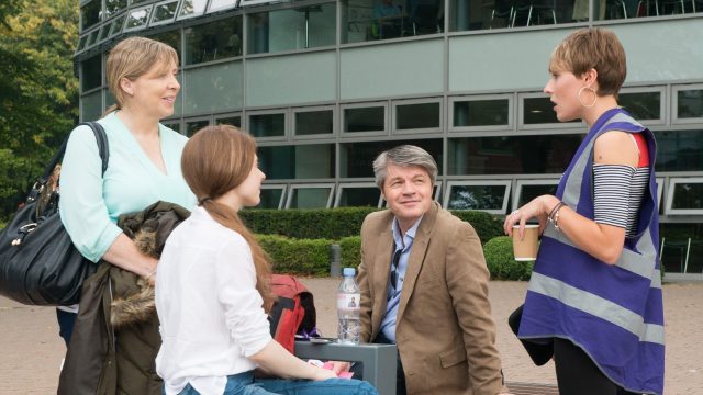 Four people are sat on a bench on the Edge Hill University campus. Three of the people are looking at the other person who is wearing a purple high-vis jacket and talking to the group of people.