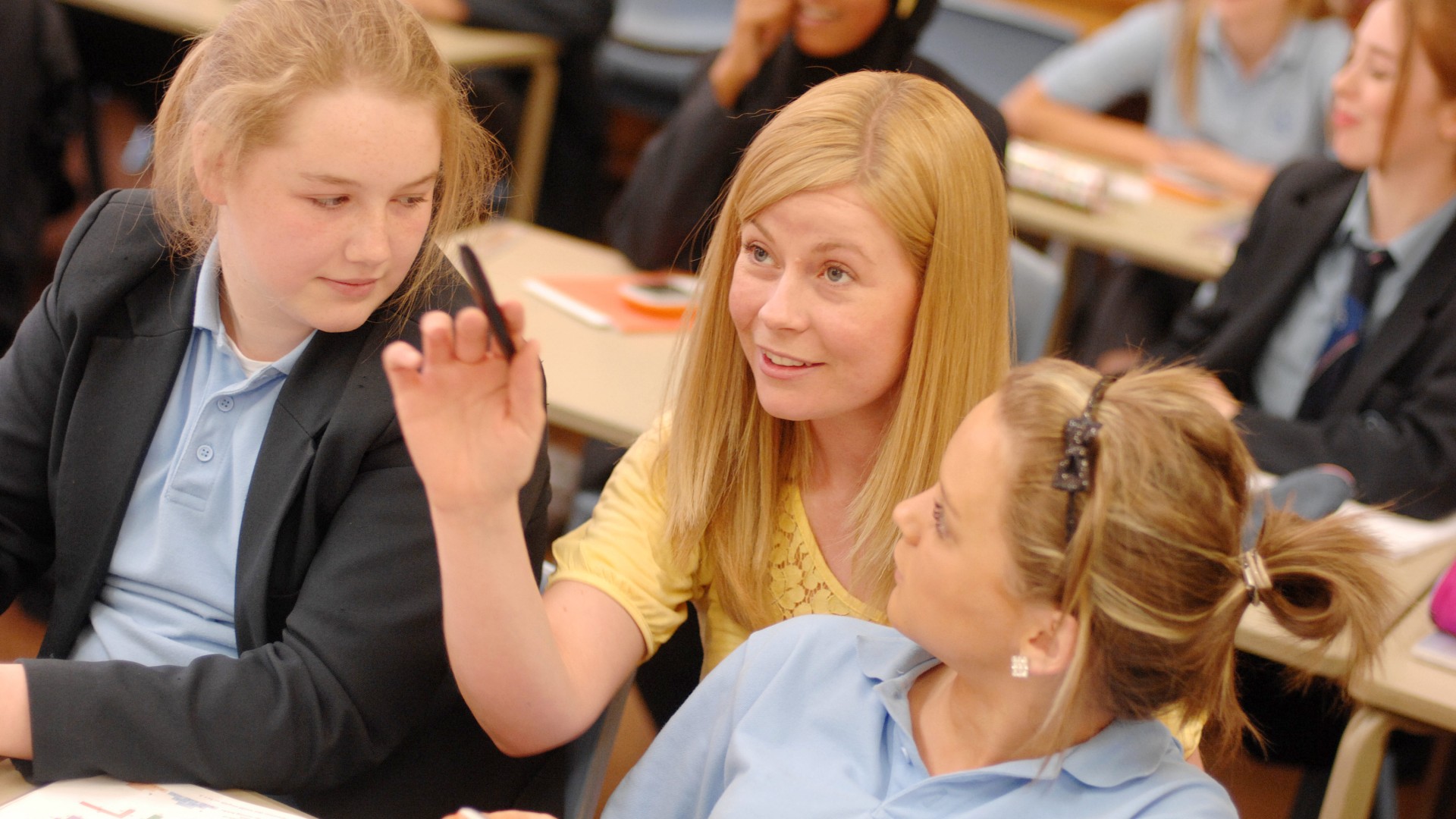 A teacher is sat in between two students pointing at something that is off-camera.