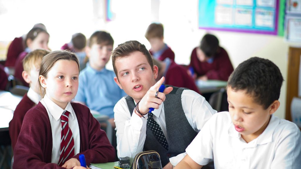 An image of a trainee teacher sat in between two students pointing towards something off-camera.