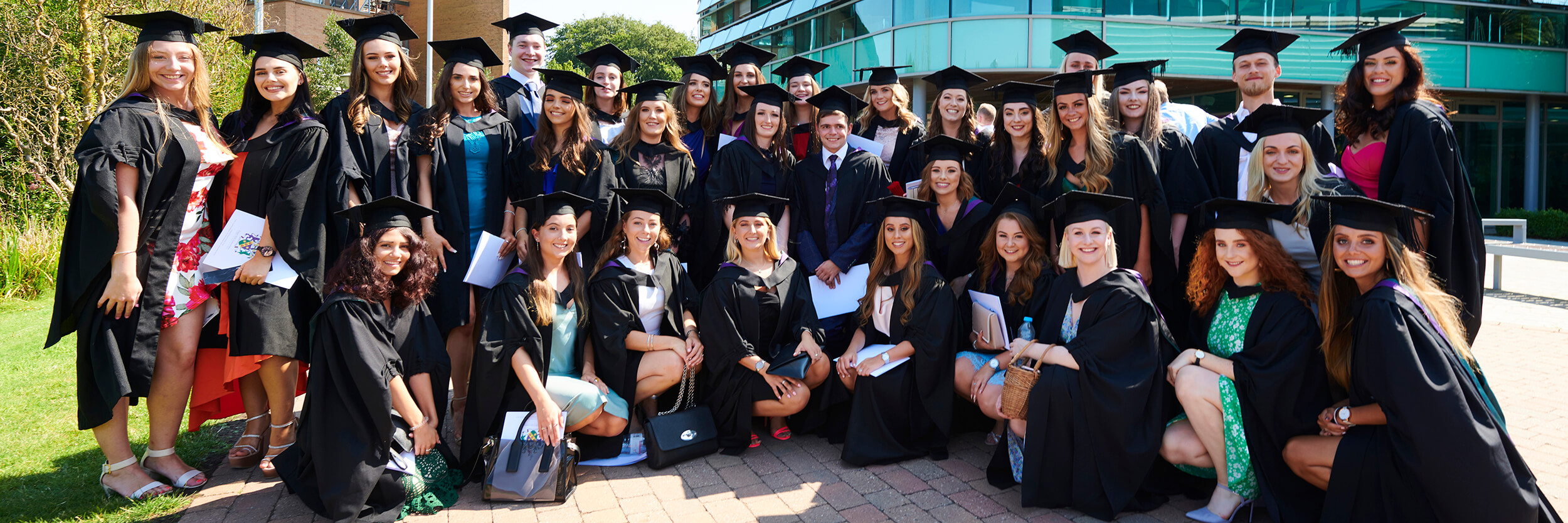 A large group of graduates pose for a photo together outside the Faculty of Health, Social Care and Medicine.