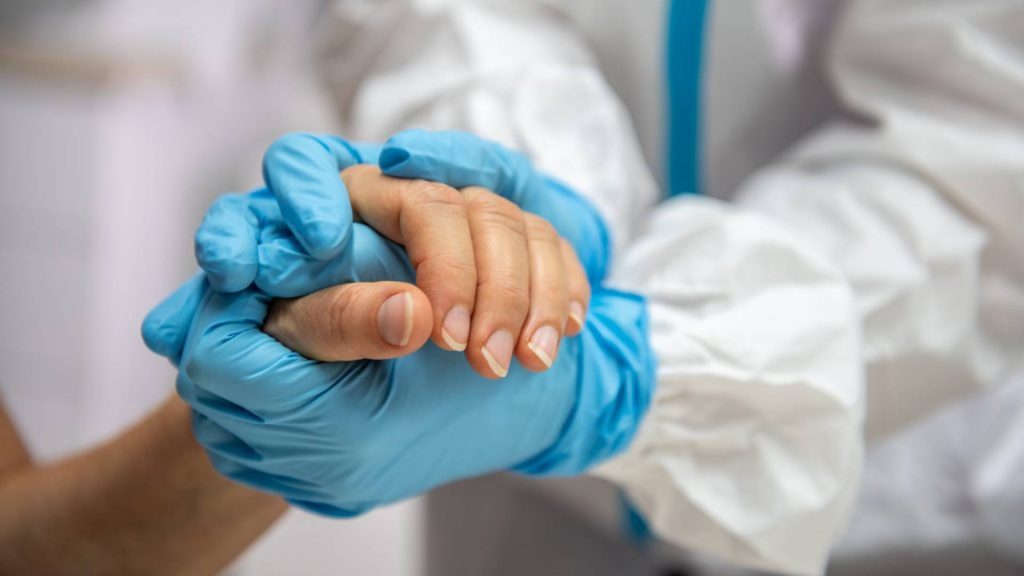 A nurse in protective clothing holds a patient's hand