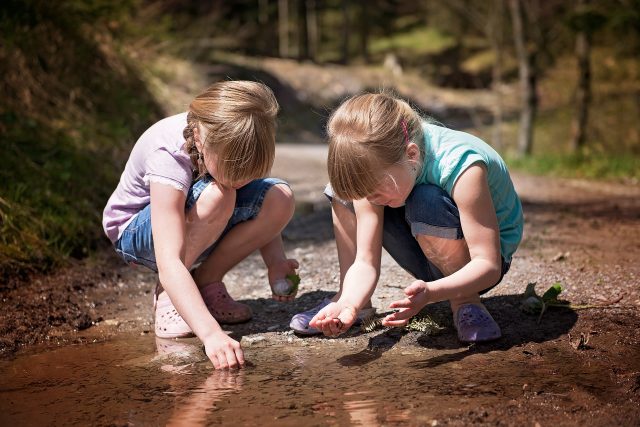 An image of two children playing with water on the ground in a forest, they are crouched down.