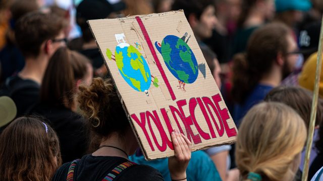 An image of multiple people attending a demonstration for climate change. The image is focusing on one person holding up a cardboard sign that says "you decide", with an image of two earths, one looking more destroyed than the other.