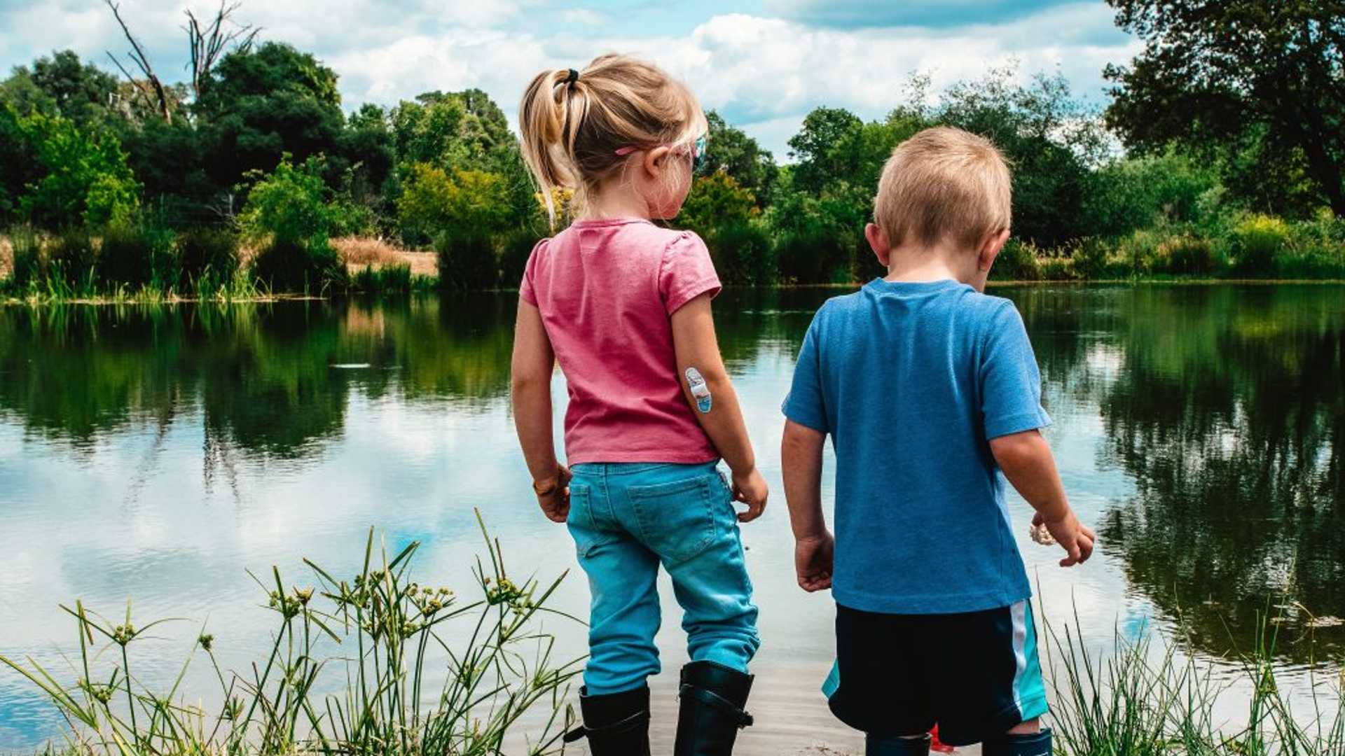 Children standing in front of a pond. They're holding hands looking into the distance.