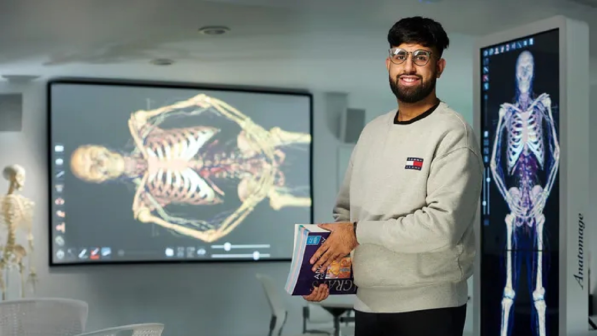 An image of a student stood in a classroom. They are stood in front of a screen on the wall that is showing an image of a skeleton. The student is holding a book. 