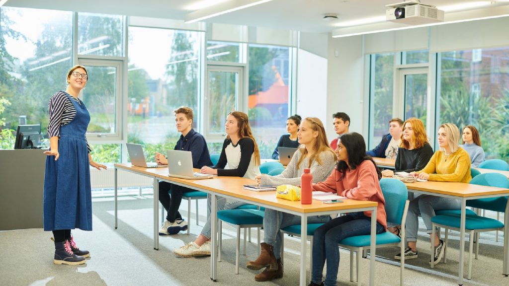 An image of students sat on three rows of desks, they are listening to a lecturer who is at the front of the room looking up at something off-camera.