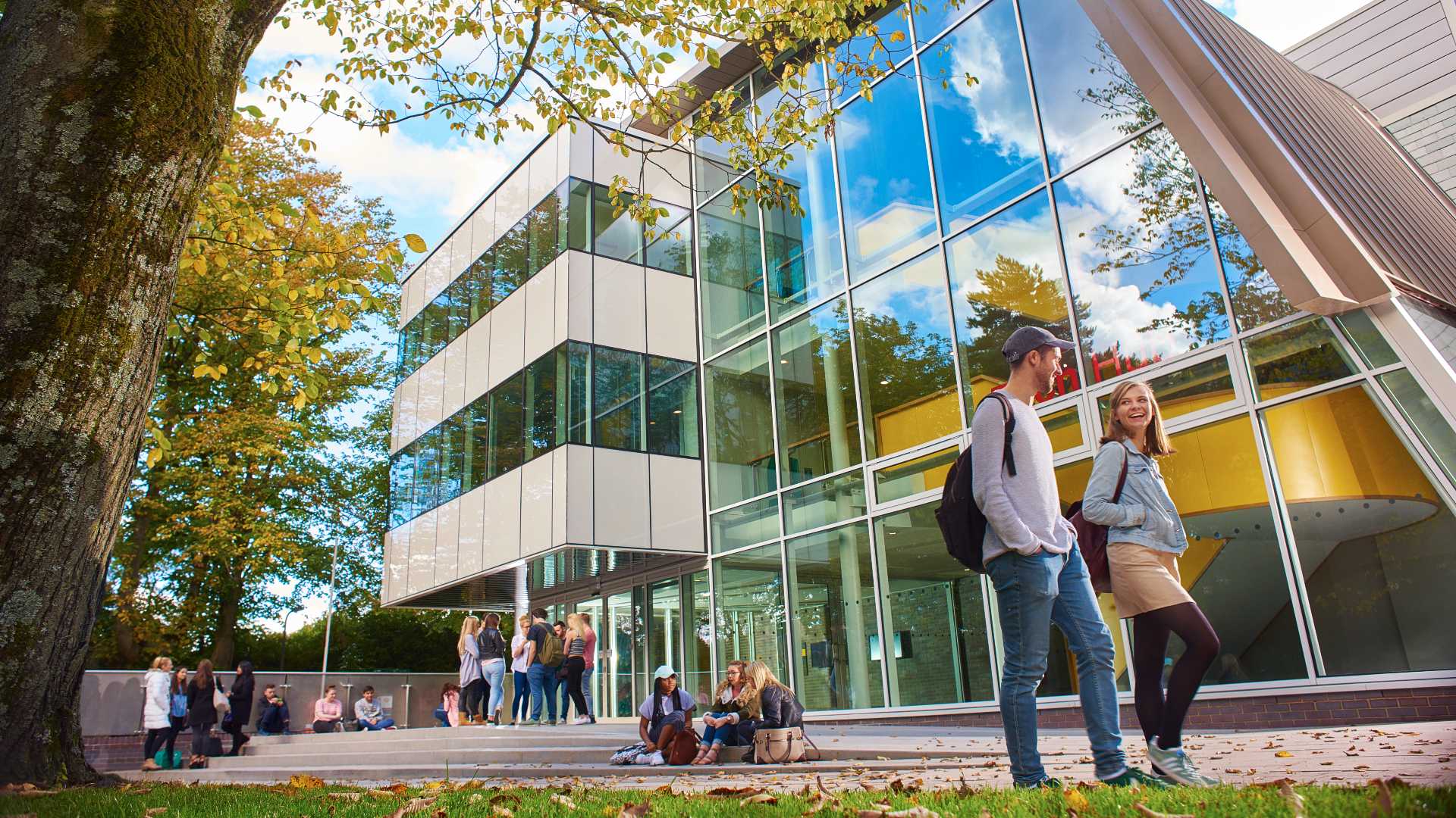 Two students walking outside tech hub building, with students sitting on steps in the background